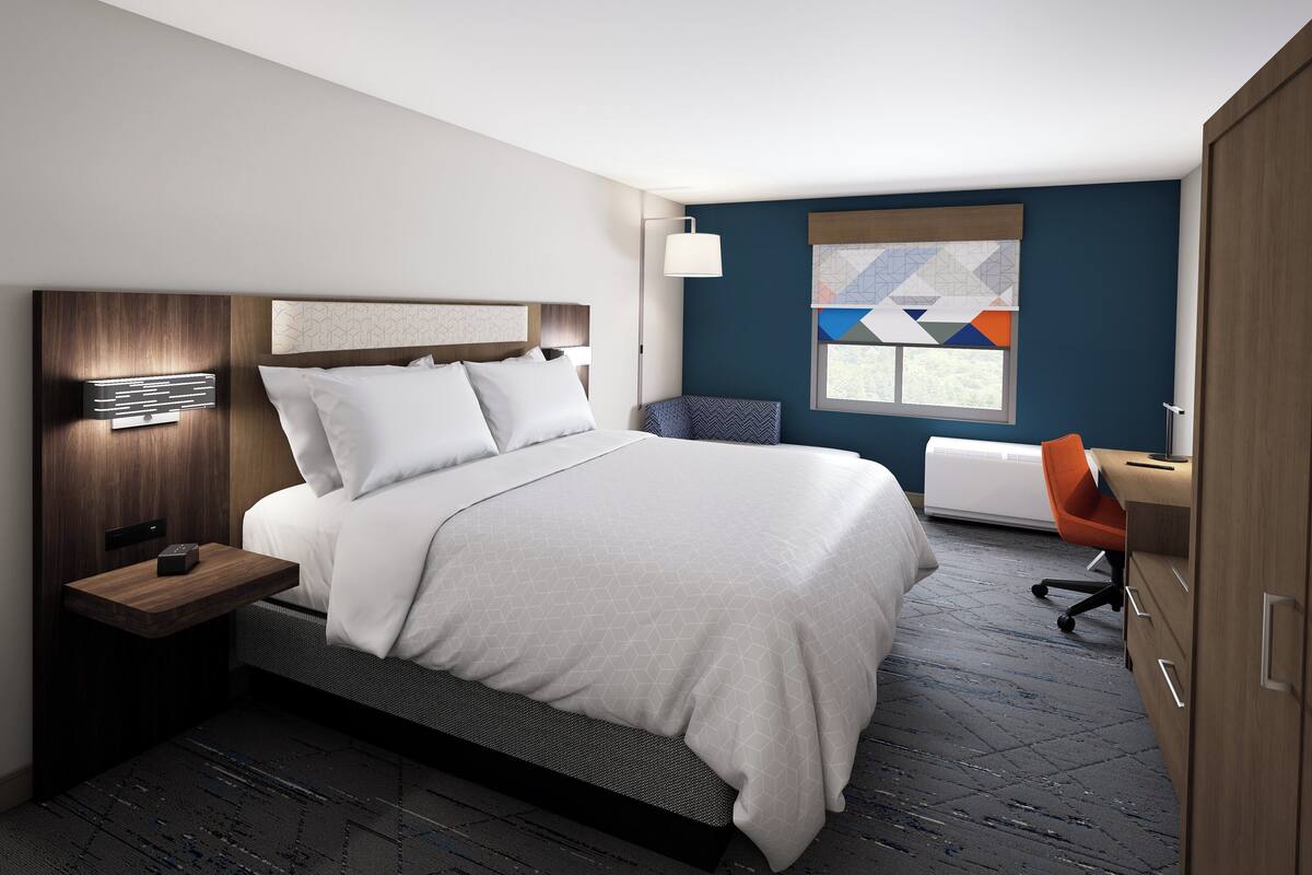 Une chambre d’hôtel moderne avec un grand lit, un bureau avec une chaise orange, une table de chevet et une lampe fixée au mur.