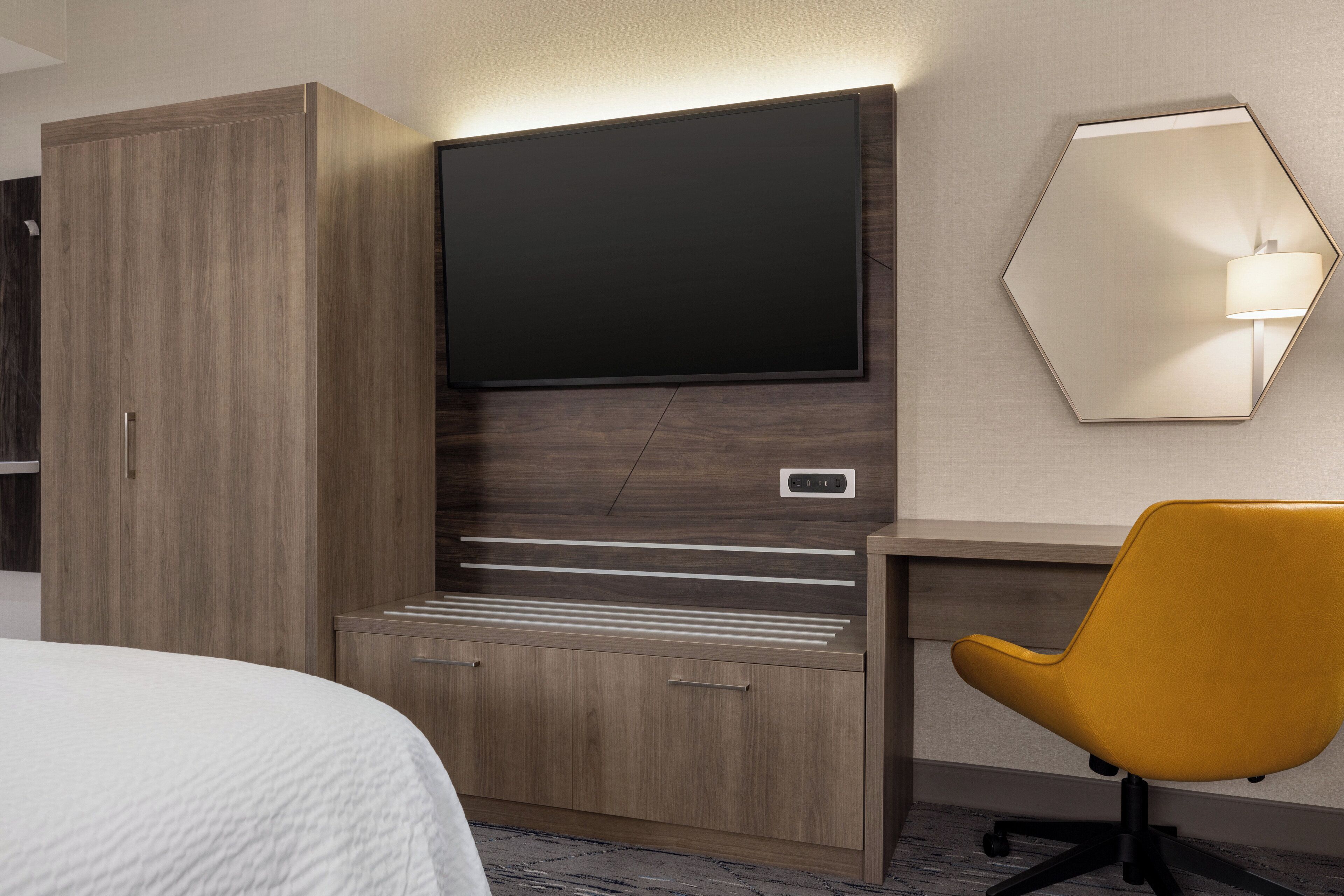 A hotel room with a bed, a wooden wardrobe, a desk with a chair, a flat-screen TV, and a wall-mounted lamp.