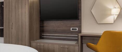 A hotel room with a bed, a wooden wardrobe, a desk with a chair, a flat-screen TV, and a wall-mounted lamp.