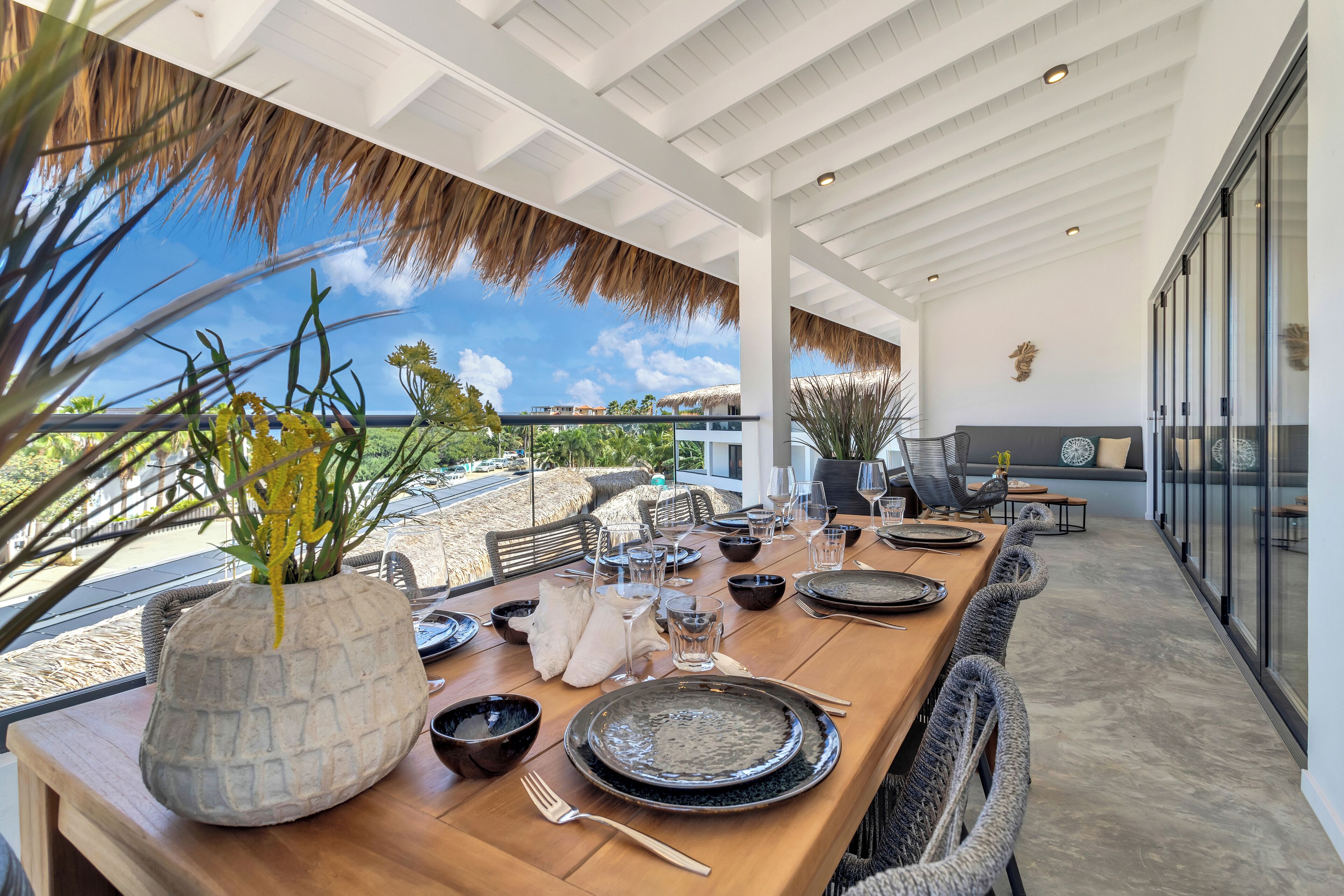 A dining area with a long wooden table set for a meal, a large decorative vase with yellow flowers, and a view of the sky and greenery outside.
