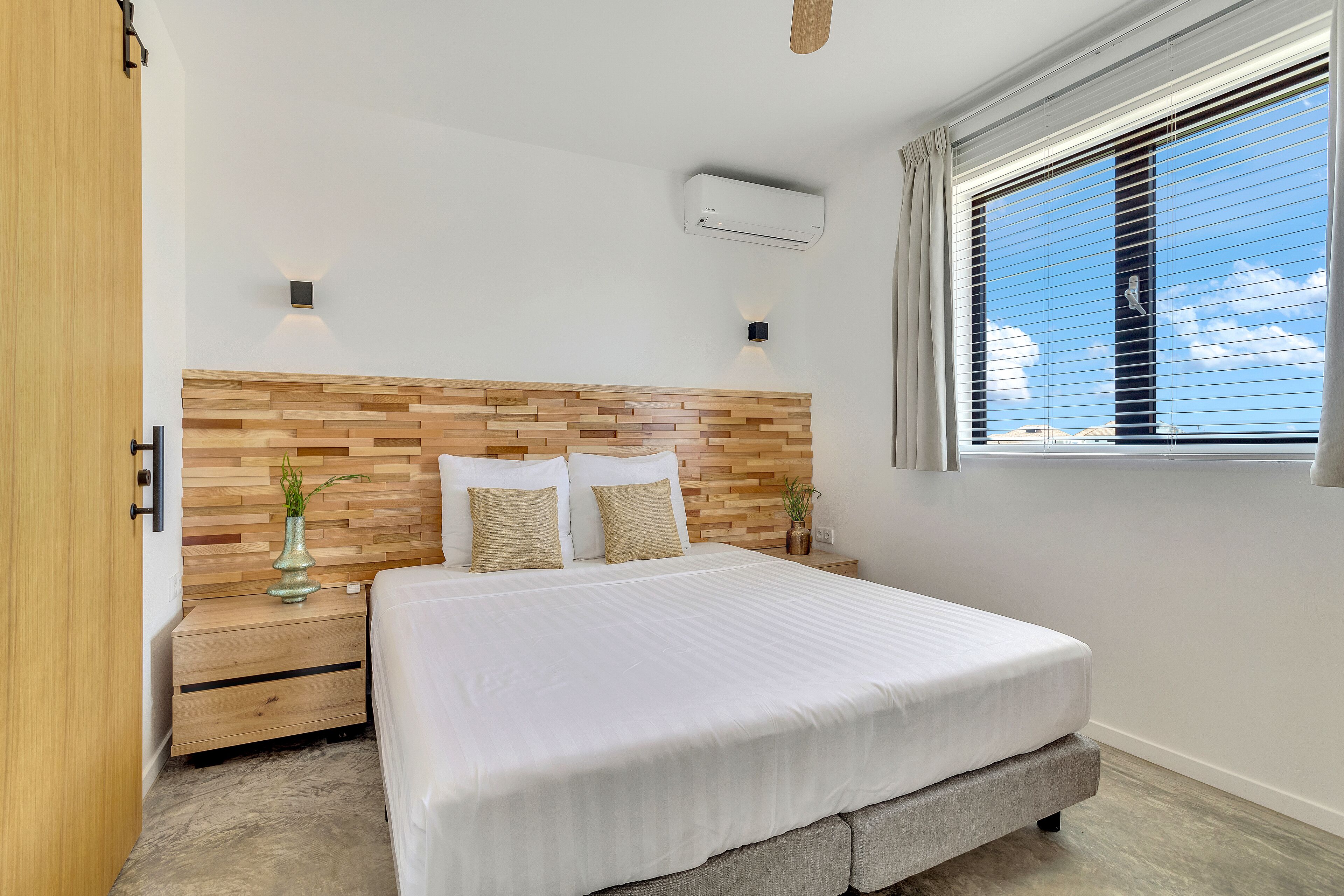 Un dormitorio con cabecera de madera, una cama grande con ropa de cama blanca, una mesita de noche con un florero y una ventana con persianas.