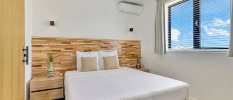 Un dormitorio con cabecera de madera, una cama grande con ropa de cama blanca, una mesita de noche con un florero y una ventana con persianas.