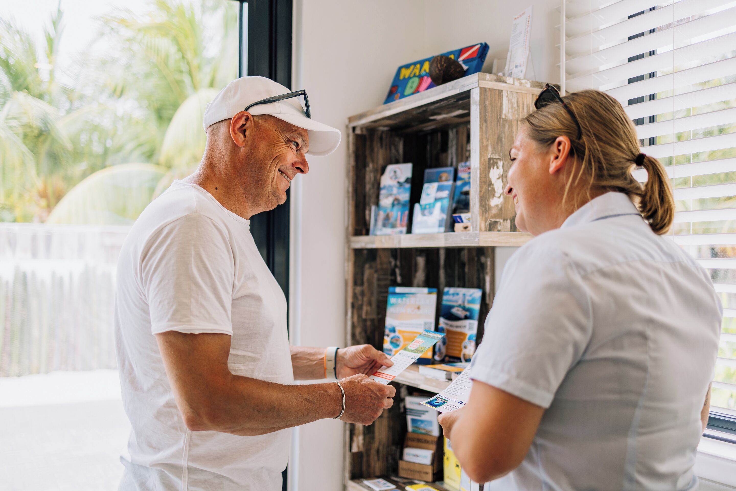 A man and a woman in a room with a window, a wooden shelf with books, and a view of palm trees outside.