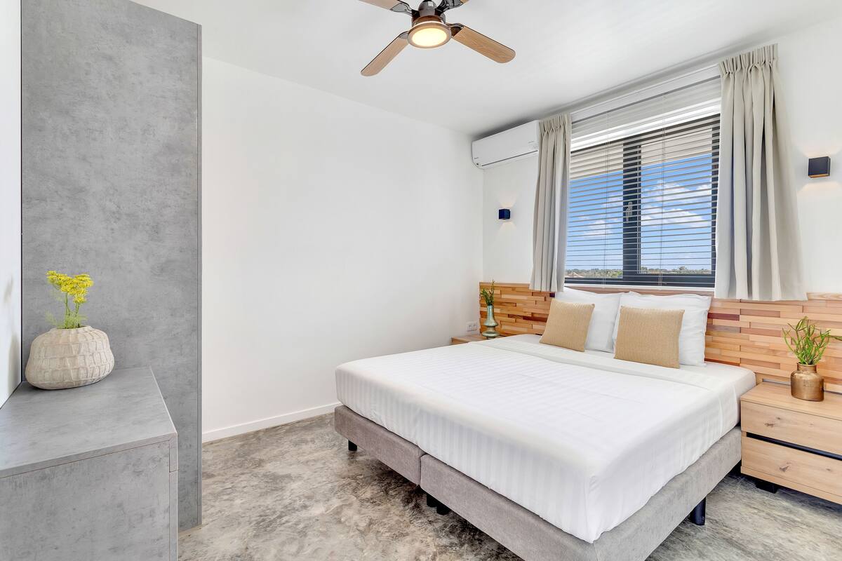 A bedroom with a bed, a nightstand, a ceiling fan, a window with curtains, and a wall-mounted air conditioner.