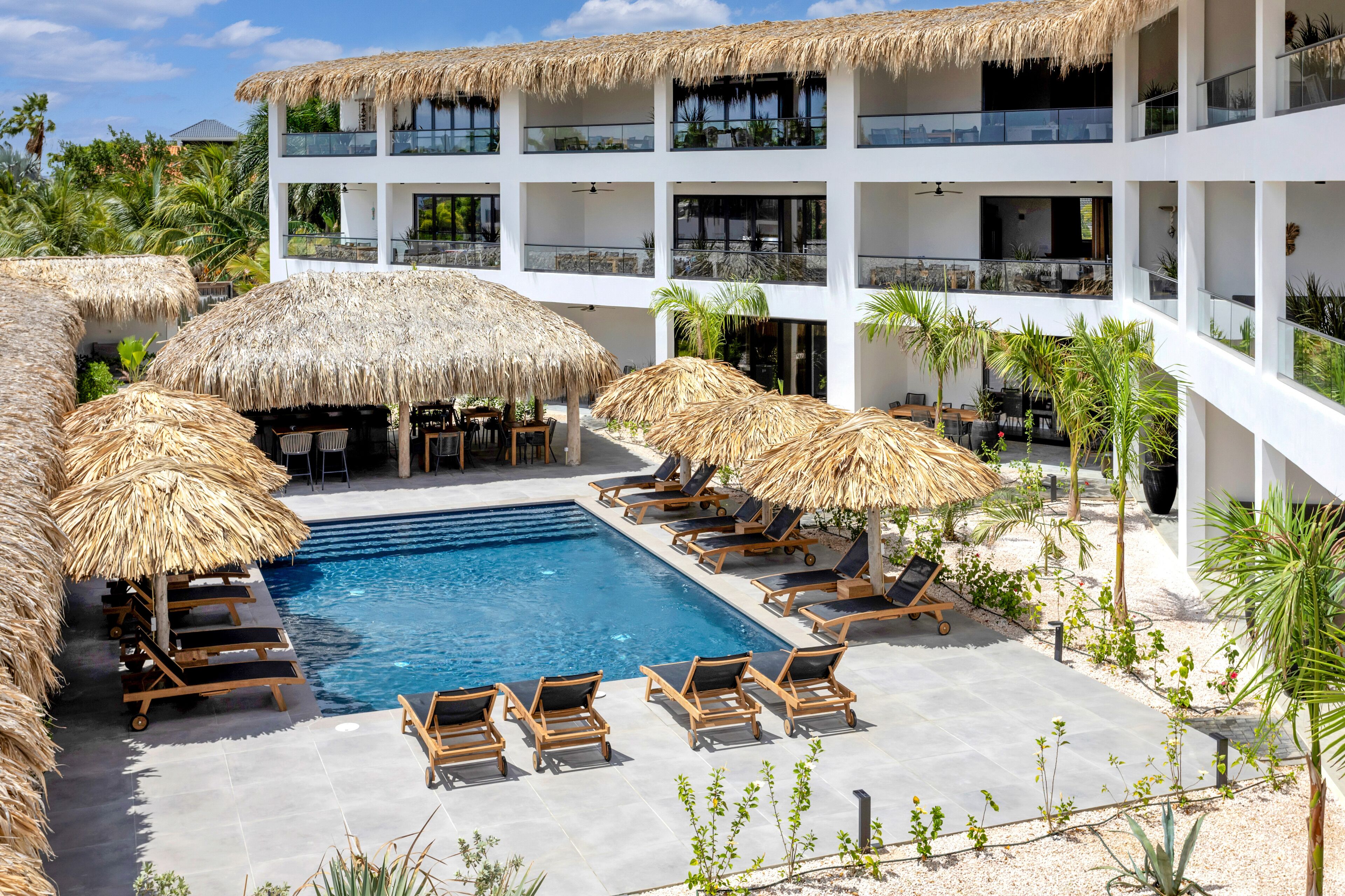 A pool area with lounge chairs and thatched umbrellas, surrounded by a white building with balconies.