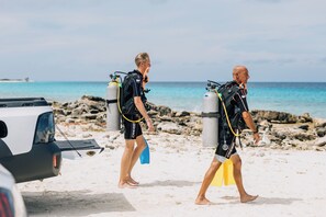 Two individuals in scuba diving gear walking on a sandy beach with rocky outcrops and the ocean in the background. - Belnem House Bonaire (Kralendijk)