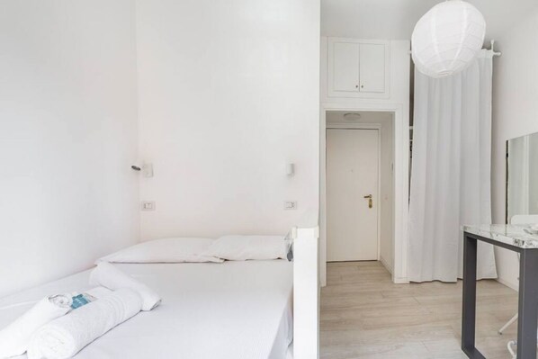 A minimalist hotel room with a single bed, a small table, and a door leading to another room.