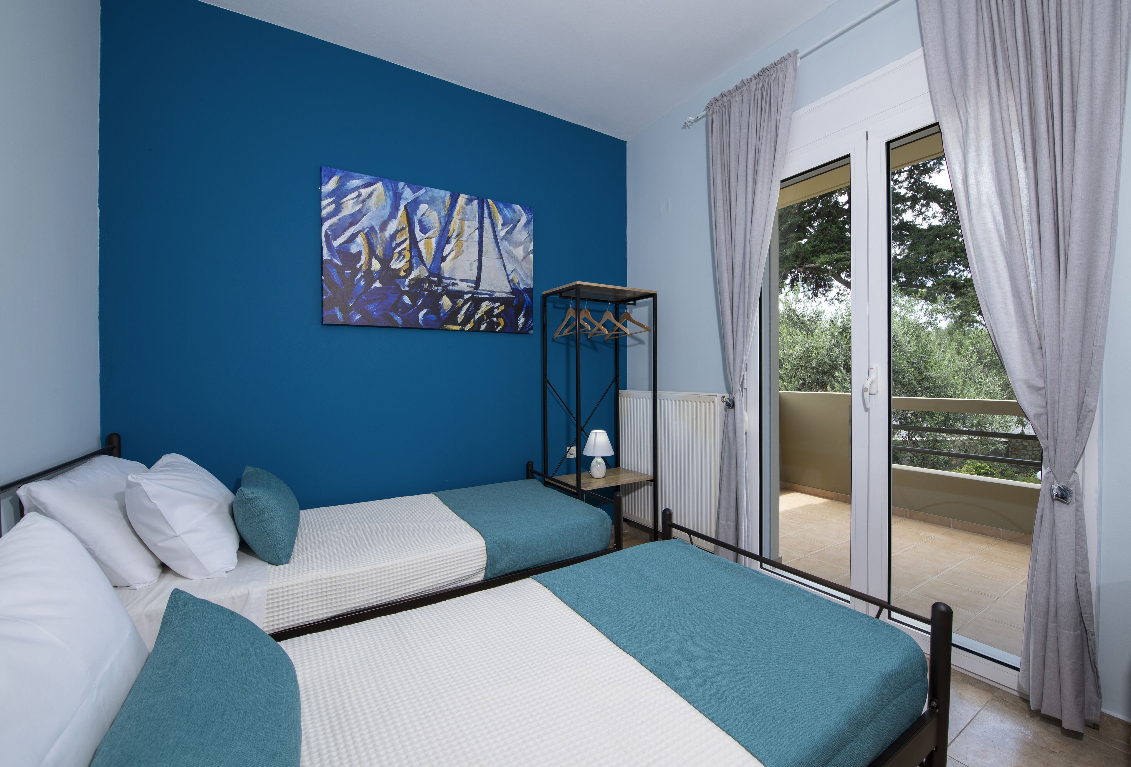 Ein Schlafzimmer mit zwei Betten, einer blauen Wand, einem Gemälde, einer Heizung und einem Balkon mit Vorhängen.