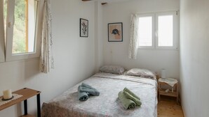 Ett sovrum med ett träpanelat tak, en säng med ett mönstrat överkast, två fönster med gardiner, ett sängbord och en liten trästol.