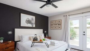 Quarto com cama, mesa de cabeceira, ventoinha no teto, obras de arte e vista através de uma porta de vidro.