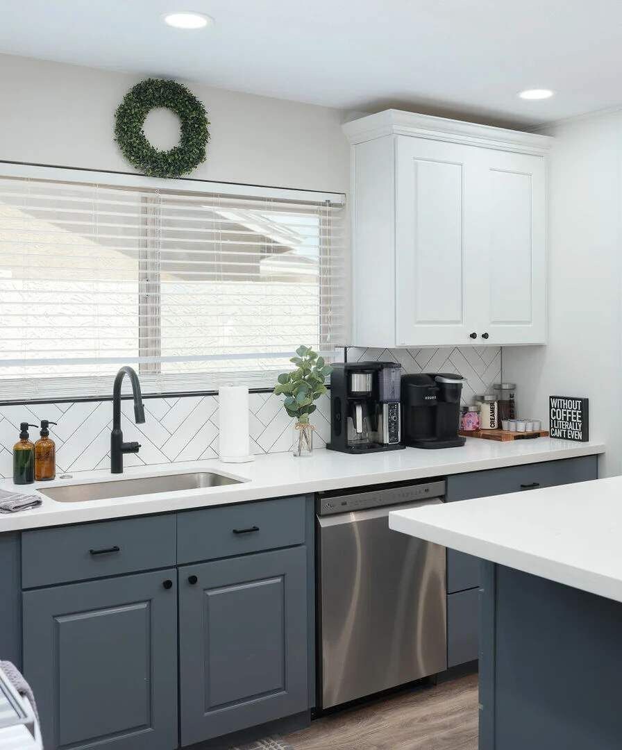 A modern kitchen with grey cabinets, a stainless steel dishwasher, a coffee maker, and a decorative wreath on the wall.