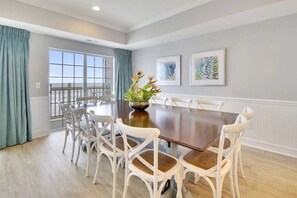 A dining room with a large table and white chairs, a window with curtains, and two framed artworks on the wall.