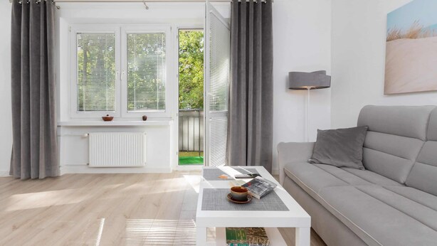 A modern living room with a white sofa, a coffee table with a book and a cup, and a view through a glass door to a garden. - Spacious Apartment | 2 Balconies | Close to the Beach (Gdańsk)