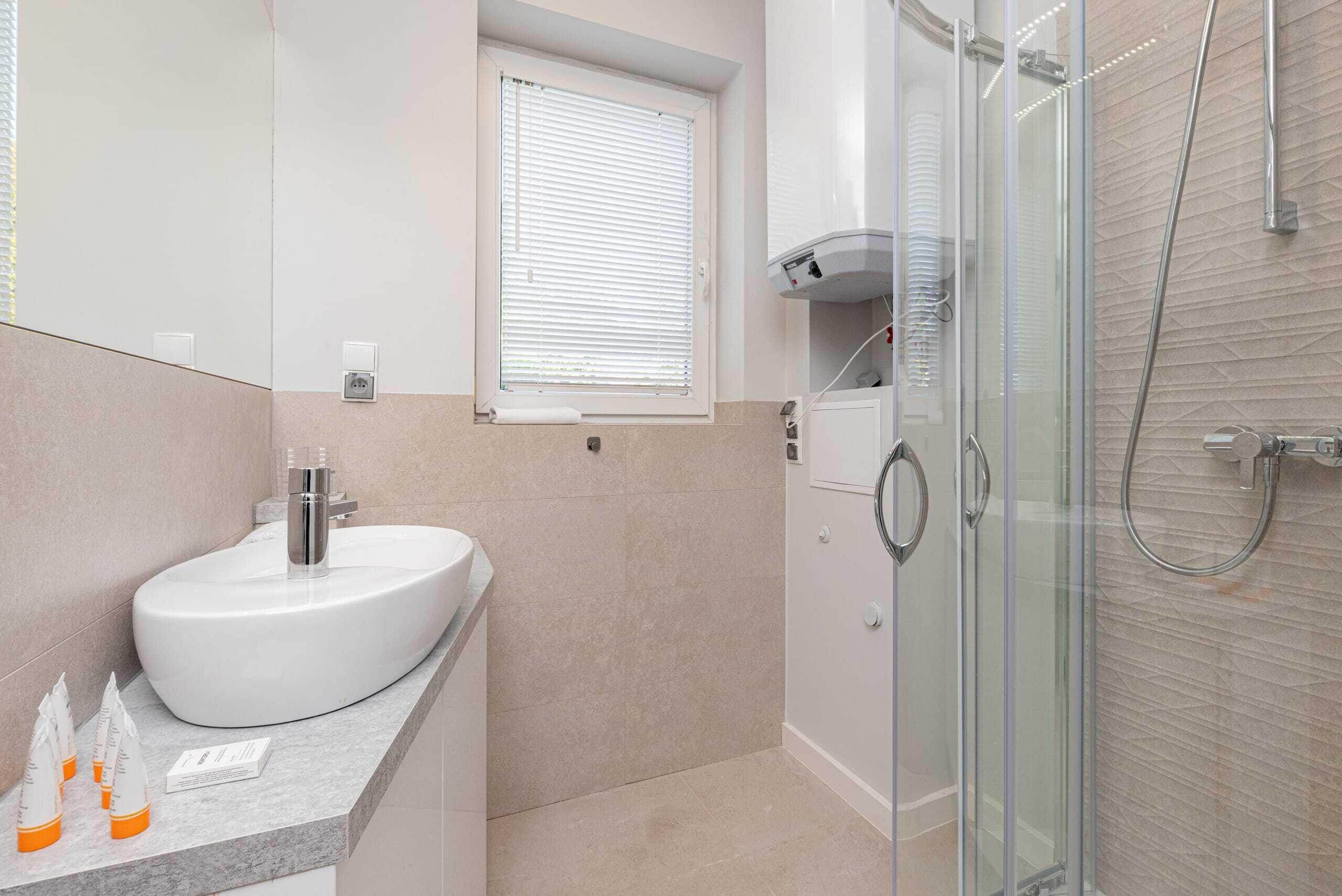 Een moderne badkamer met een wit wastafelmeubel, een douchecabine van glas en een raam met zonwering.