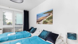 A bedroom with two beds, a window with blinds, a radiator, a painting of a beach, and a bedside lamp. - Spacious Apartment | 2 Balconies | Close to the Beach (Gdańsk)