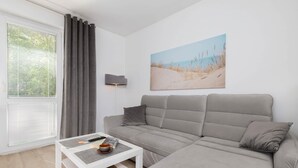 A modern living room with a grey sofa, a white coffee table, and a painting of a beach scene. - Spacious Apartment | 2 Balconies | Close to the Beach (Gdańsk)