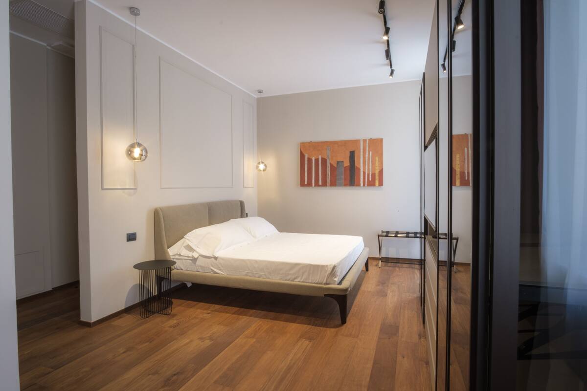 A modern bedroom with a bed, a small side table, and a wall-mounted light fixture.