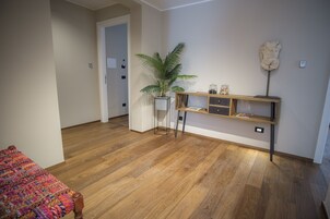 Ein Zimmer mit Holzboden, einem Bett mit bunter Decke, einer kleinen Pflanze, einem Regal mit Dekoration und einer Tür zu einem anderen Raum.