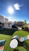 Ein modernes Haus mit Flachdach, kleinem Hinterhof mit Kunstrasen und einer Terrasse mit zwei Liegestühlen und einem kleinen Tisch.