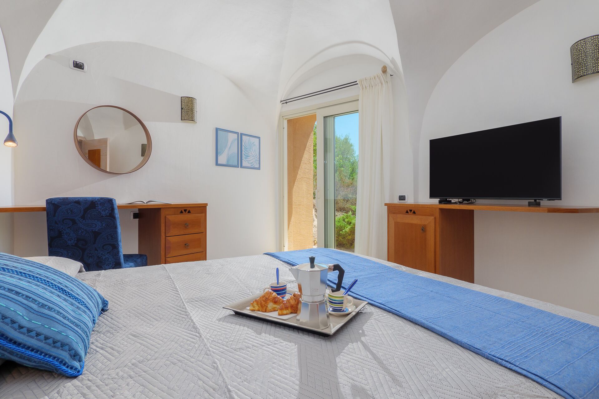 A hotel room with a bed, a desk, a chair, a television, and a window with a view of greenery.
