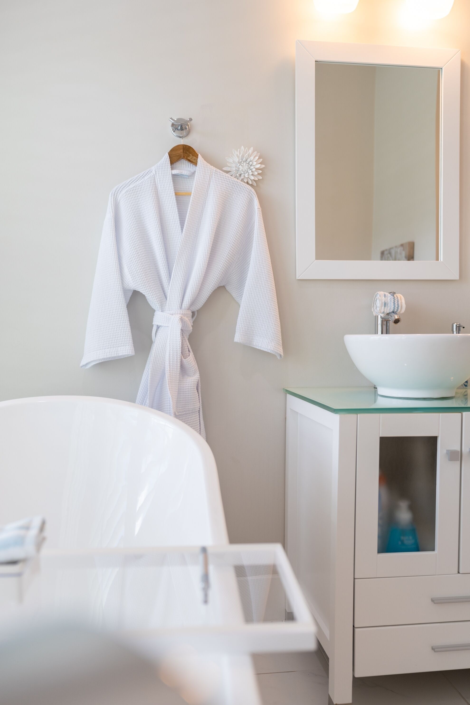 A bathroom with a white bathtub, a glass sink, and a white robe hanging on the wall.