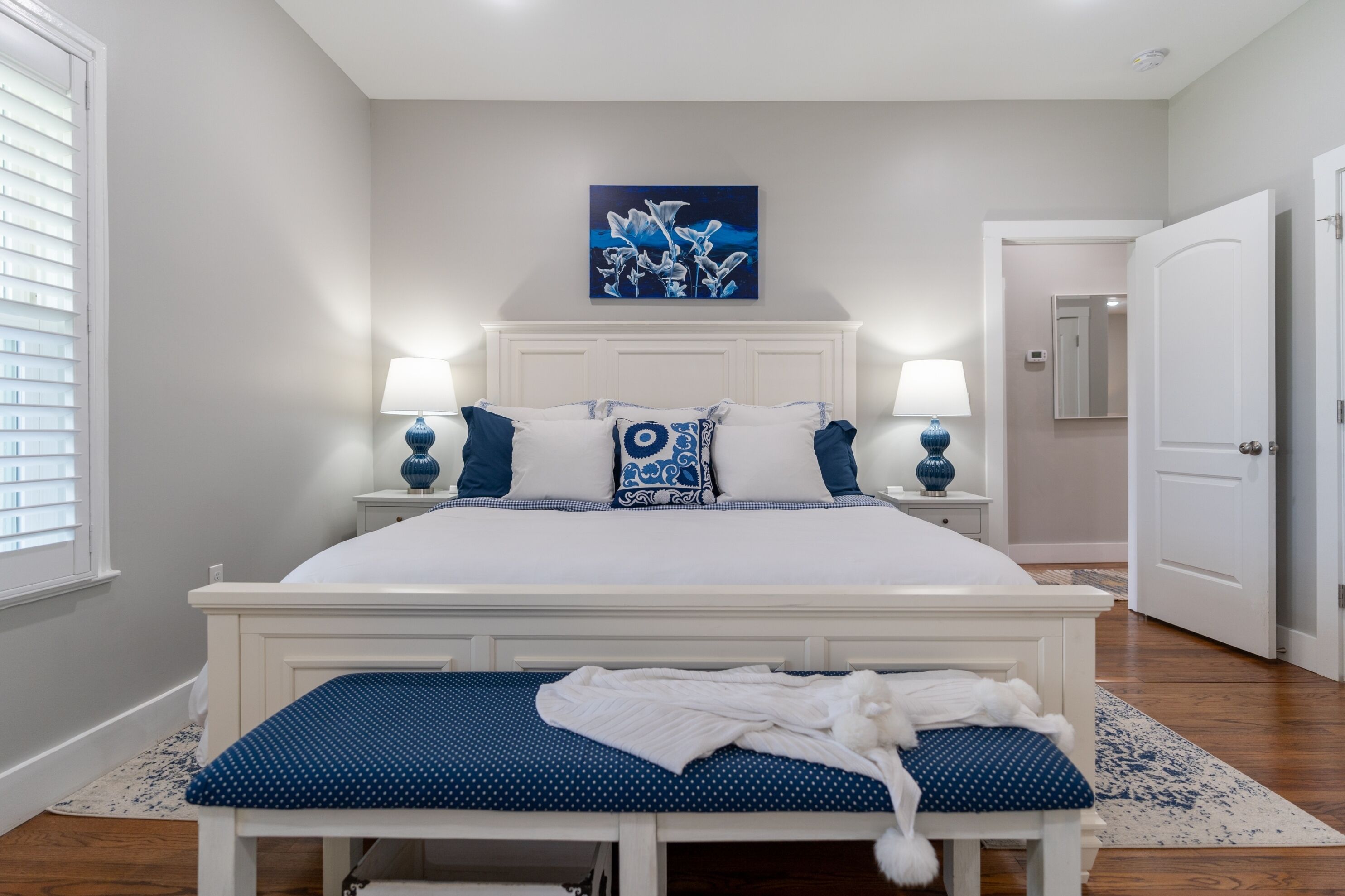 Une chambre avec un grand lit, deux tables de chevet avec des lampes, un banc aux motifs bleus et blancs, et un tableau accroché au mur.
