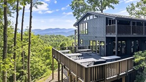Una casa moderna de dos pisos con una terraza, rodeada por un bosque y montañas al fondo.