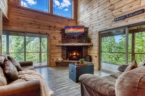 A cozy cabin interior with a fireplace, leather sofas, and a large window offering a view of the forest.