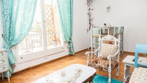 Um quarto com piso de madeira, uma mesa branca com toalha branca, uma poltrona estampada e um tapete azul.