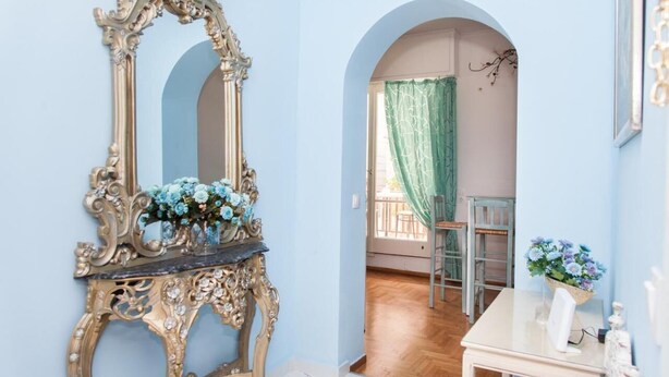 Um quarto com um espelho grande e decorativo, uma pequena mesa com um vaso de flores, e vista através de uma porta arqueada para outro quarto com uma cortina verde.