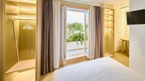 A hotel room with a bed, a TV, a desk, and a view of greenery outside. - Hotel Leonor (Conil de la Frontera)