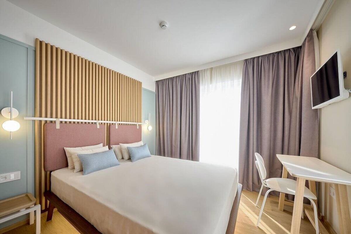 A hotel room with a bed, a desk, a chair, a TV, and a window with curtains.