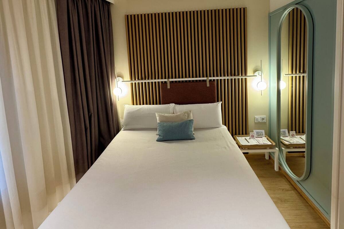 A hotel room with a large bed, a headboard with vertical slats, a bedside table with a lamp, a mirror, and a wooden floor.