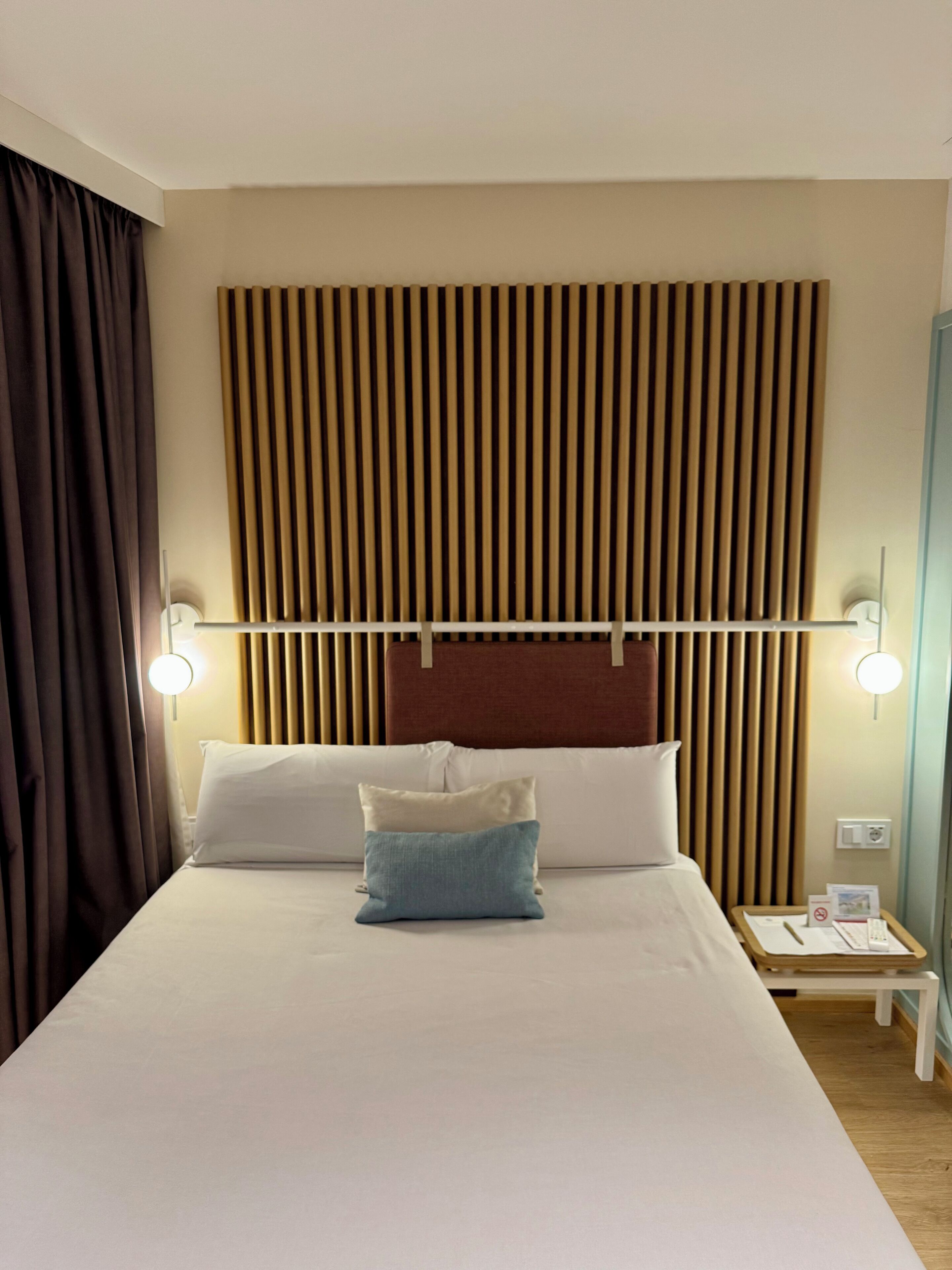 Ein Hotelzimmer mit einem großen Bett, zwei Nachttischen, einem Holzkopfende und einem Vorhang auf der linken Seite.