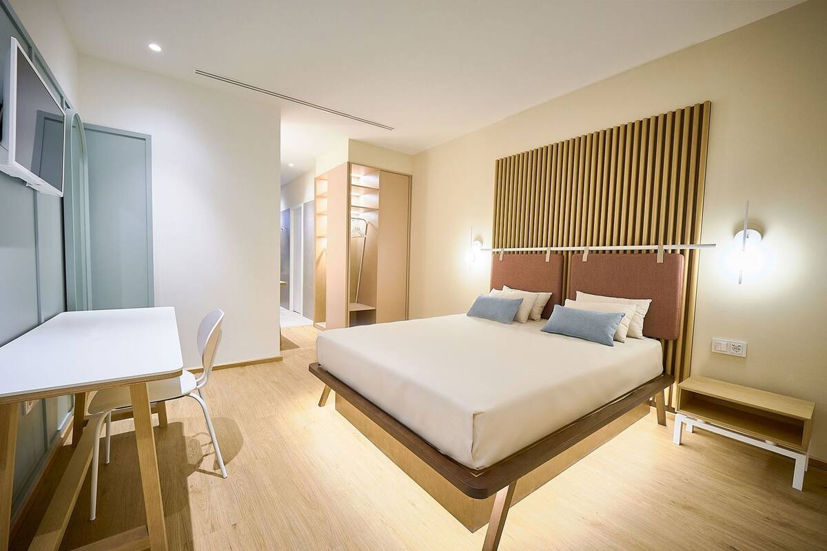 A modern hotel room with a bed, bedside table, desk, chair, and a wardrobe.