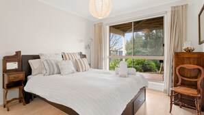 A bedroom with a large bed, wooden nightstands, a wooden chair, and a window with a view of greenery.