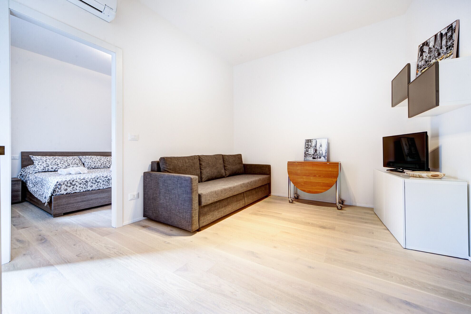 Ein modernes, minimalistisches Zimmer mit einem Bett, einer Couch, einem kleinen Holztisch und einem Fernseher.