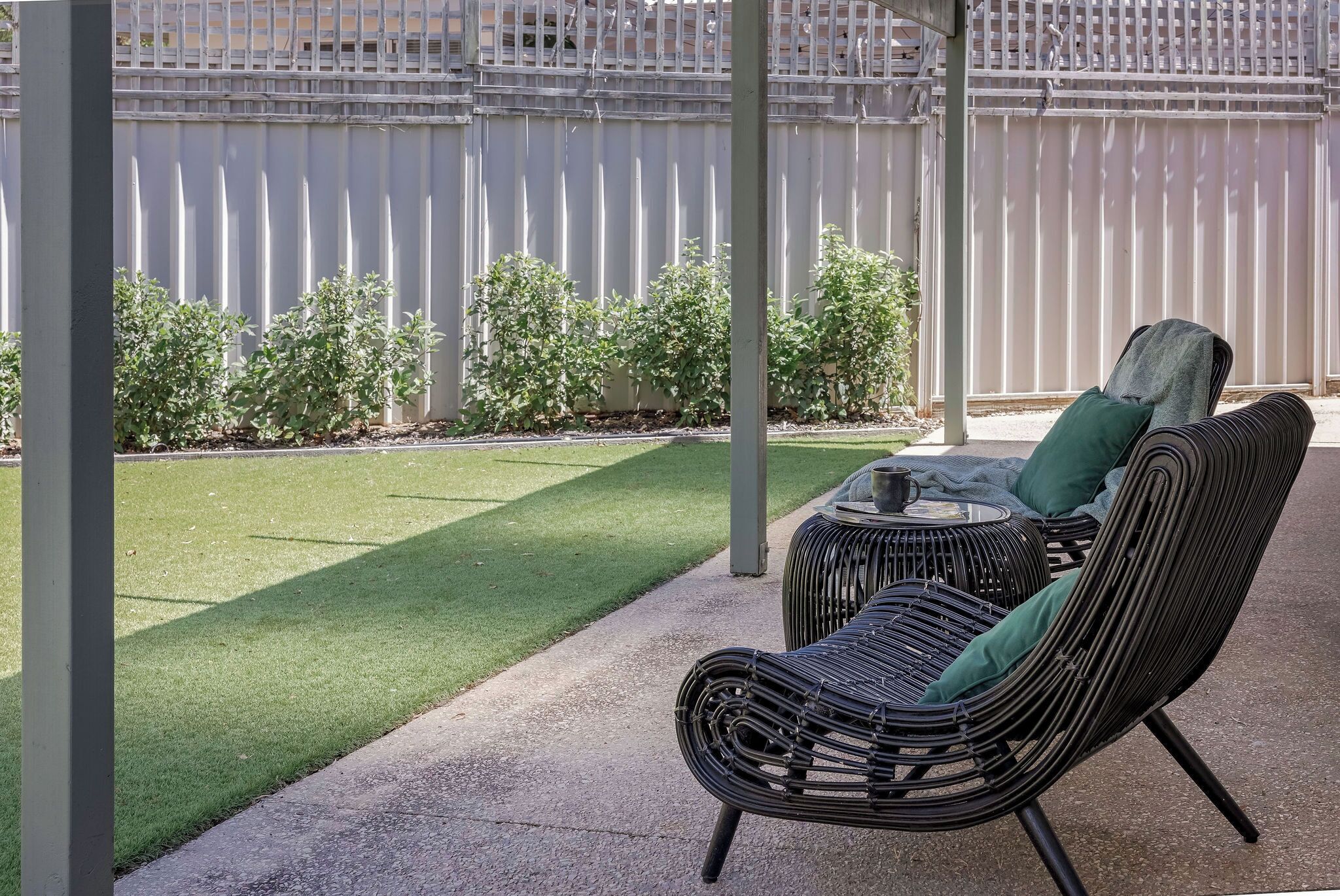 A covered patio area with a wicker chair and a small table, a neatly trimmed lawn, and a white fence in the background.
