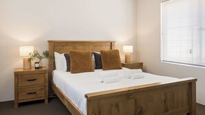 A neatly made bed with white linens and brown pillows, a wooden headboard, two bedside tables with lamps, and a window with blinds. - GORGEOUS & WELCOMING COASTAL COTTAGE CLOSE TO TOWN (Dunsborough)