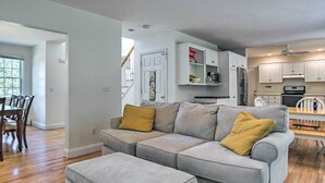 Ein modernes Wohnzimmer mit einem grauen Ecksofa, einem Essbereich mit Holztisch und -stühlen sowie einer Küche mit weißen Schränken und Geräten.