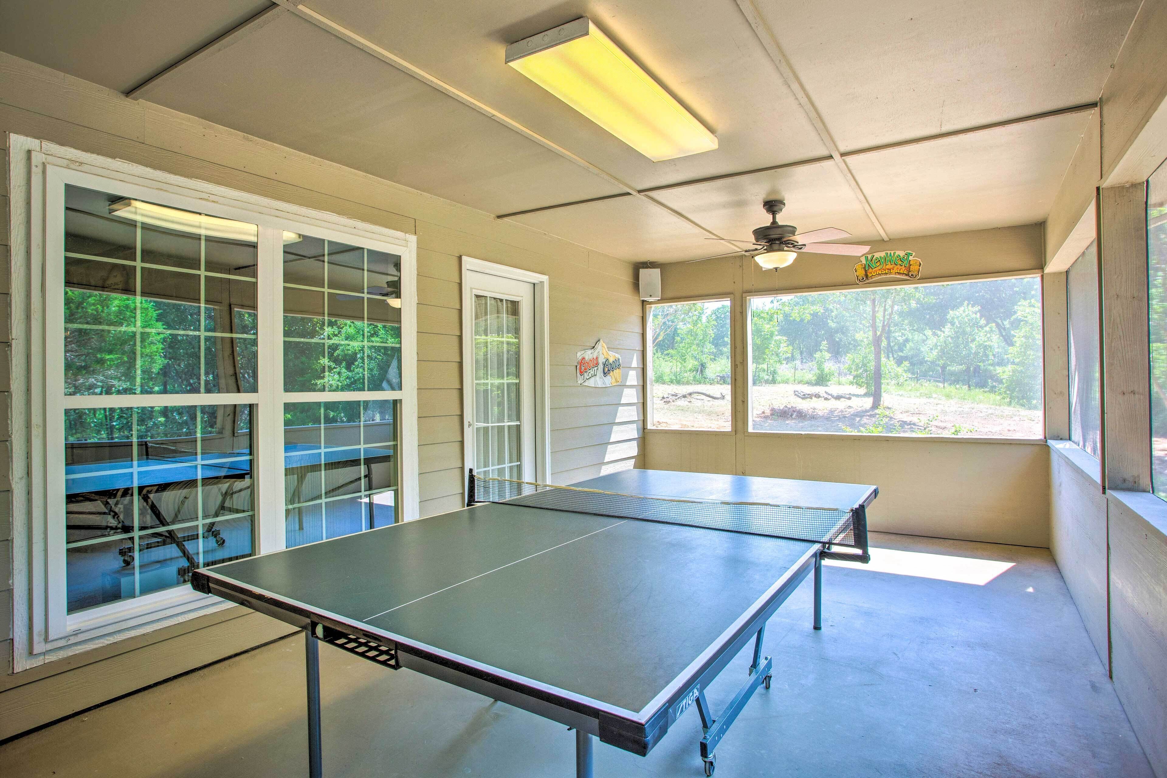 Una mesa de ping pong en una habitación con ventanas amplias, un ventilador de techo y un cartel sobre las ventanas.