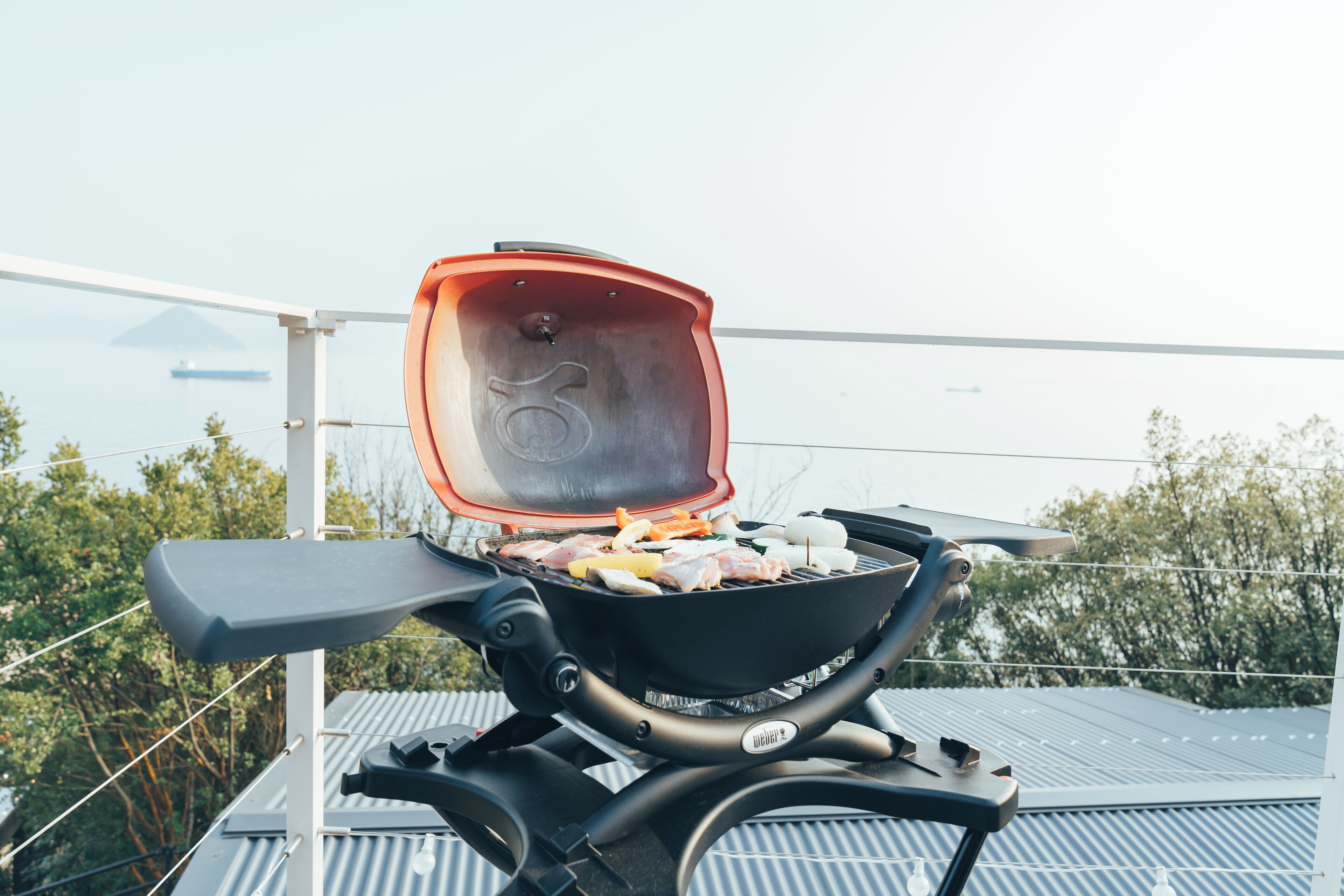 Una parrilla portátil con comida cocinándose, montada en un balcón con vista a árboles y un barco a lo lejos.