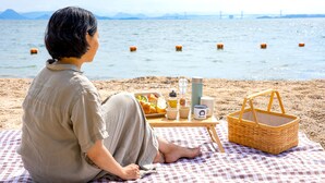 Una persona sentada sobre una manta de cuadros junto al agua, con una preparación para picnic que incluye una cesta, comida y bebidas.