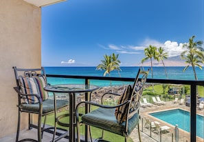 A balcony with two chairs and a table, overlooking a pool and a view of the ocean with palm trees and a mountain in the distance.