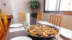 Une table à manger avec un grand plat de paella aux fruits de mer, des assiettes blanches et un verre orné d’un motif coloré.