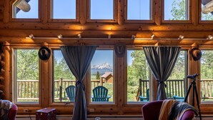 A log cabin interior with large windows, a wooden ceiling, a table with books and a glass, and a chair with a blanket. - Beautiful Moran Home with spectacular Teton Views (Moran)