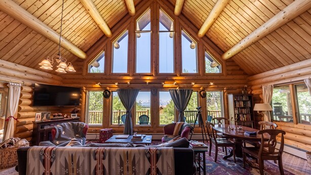 A log cabin interior with a high ceiling, large windows, a cozy seating area, and a dining table. - Beautiful Moran Home with spectacular Teton Views (Moran)