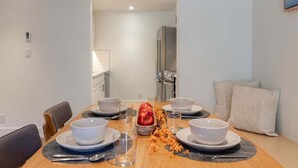 Un coin repas avec une table en bois dressée pour quatre personnes, des assiettes blanches, des bols et des couverts. Une pomme rouge et un bol de noix sont posés sur la table.