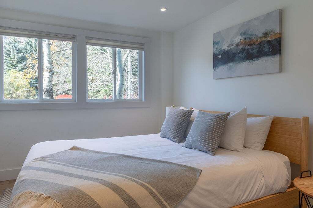 A bedroom with a bed, pillows, a blanket, a painting, and a window with a view of trees.