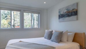 Une chambre à coucher avec un lit, des oreillers, une couverture, un tableau et une fenêtre donnant sur des arbres.
